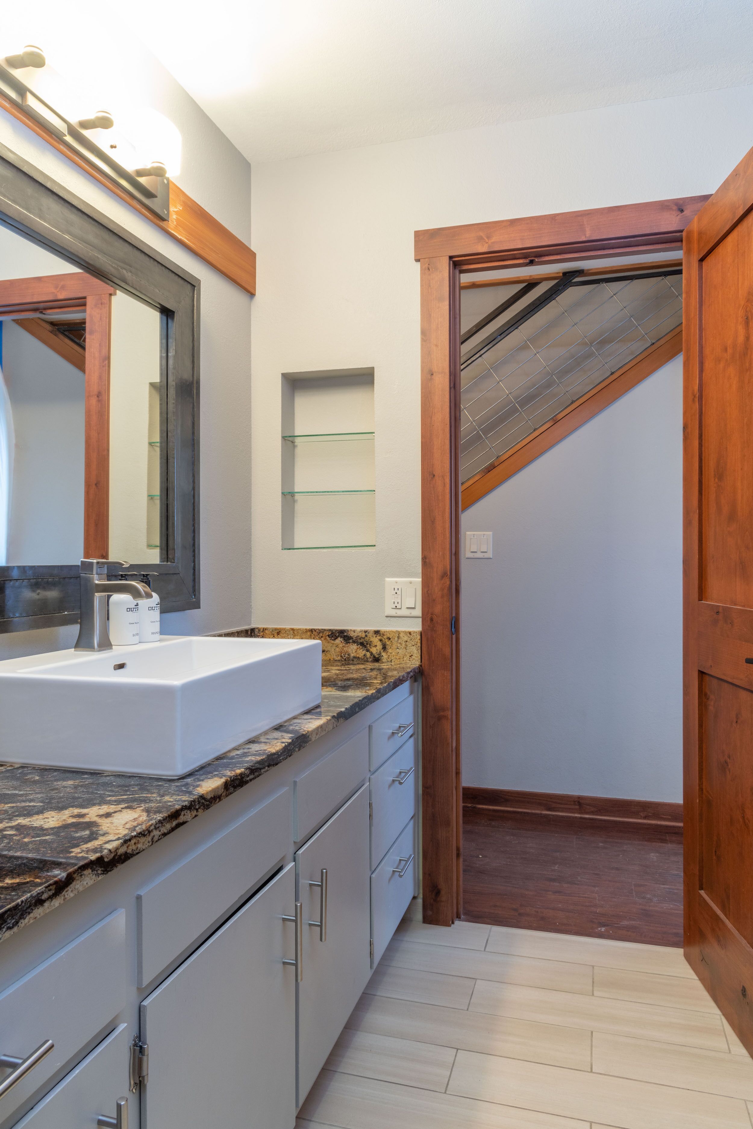 Un baño moderno con encimera de granito, un espejo grande y una puerta de madera que da a la zona de la ducha.