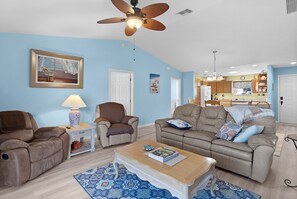 A living room with a ceiling fan, a sofa, a coffee table, and a dining area with a kitchen in the background. - Ranch Retreat: Luxury Living in Village Charm! (The Villages)