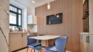 Een moderne keuken met een ronde tafel, blauwe stoelen, een aan een houten wand bevestigde televisie, en een raam dat natuurlijk licht binnenlaat.
