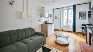 A modern apartment with a green sofa, a small dining table, a kitchen with white cabinets, and a large window with a view. - 21.Studio 2Pers#Sèvres-Lecourbe#Champ-de-Mars (Paris)