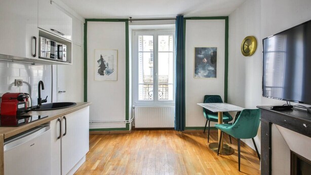 A compact kitchen with white cabinets, a red coffee machine, a window with a view, a table with two chairs, and a TV on the right. - 21.Studio 2Pers#Sèvres-Lecourbe#Champ-de-Mars (Paris)