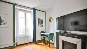 A modern living room with a fireplace, a dining area with two chairs, a TV mounted on the wall, and a large window with curtains. - 21.Studio 2Pers#Sèvres-Lecourbe#Champ-de-Mars (Paris)