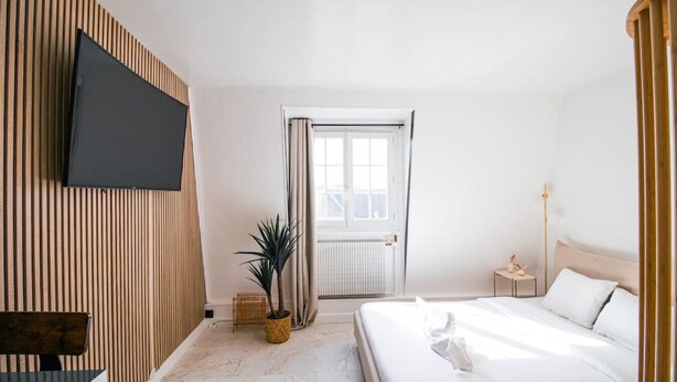 A modern hotel room with a large bed, a flat-screen TV mounted on the wall, a potted plant, and a window with curtains. - Typical Parisian Studio in Le Marais (Paris)