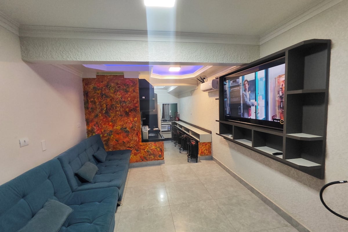 Una sala de estar con un sofá azul, un televisor de pantalla plana montado en la pared y una cocina al fondo.