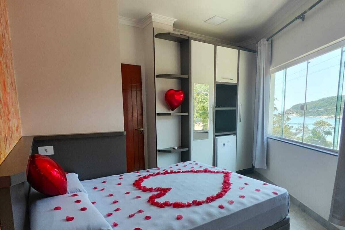 Un dormitorio moderno con una cama decorada con pétalos de rosa formando un corazón, una decoración de corazón rojo en la pared y vistas a un paisaje costero a través de amplias ventanas.