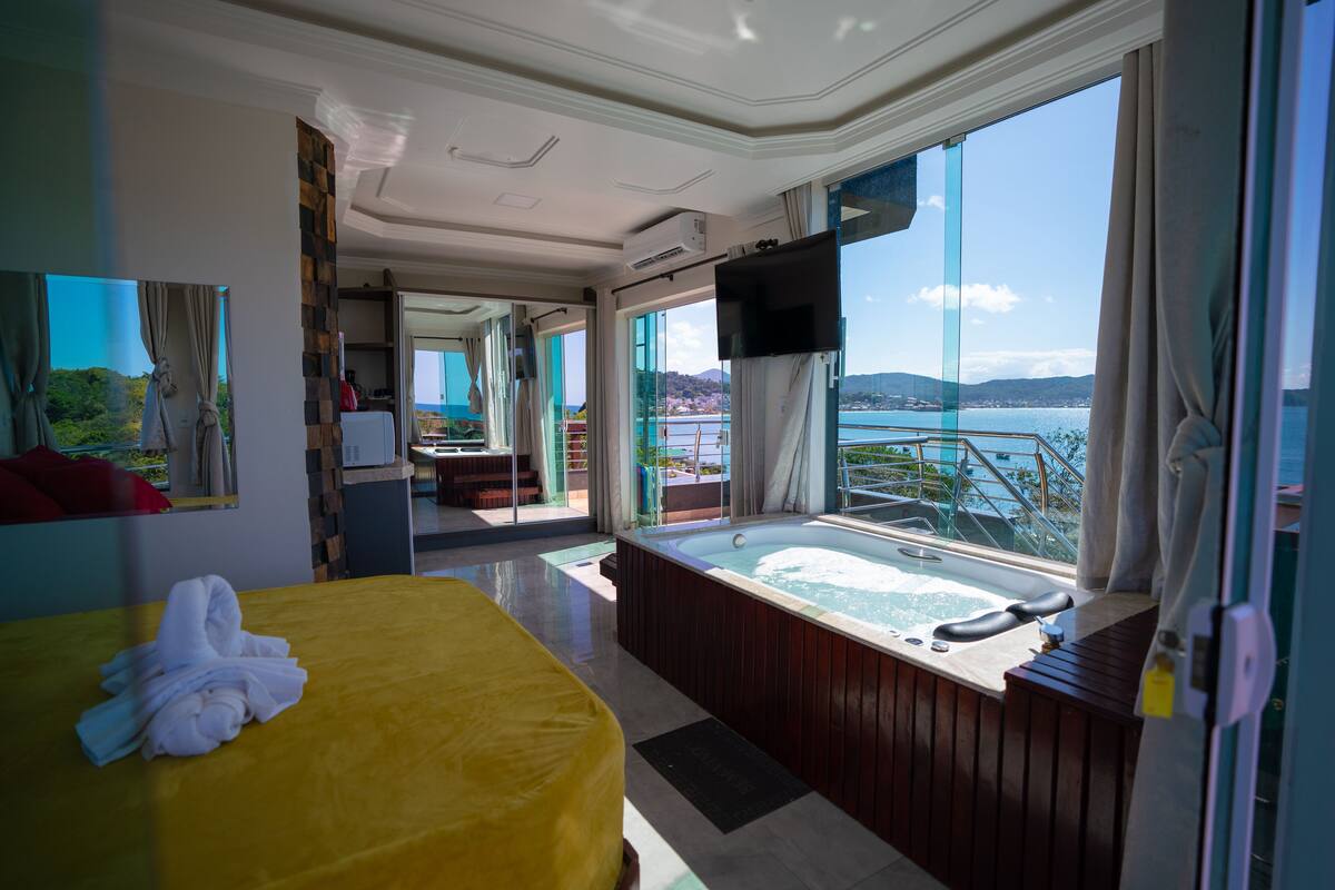 Habitación de hotel con una cama grande, una bañera, un televisor y un balcón con vista al mar.