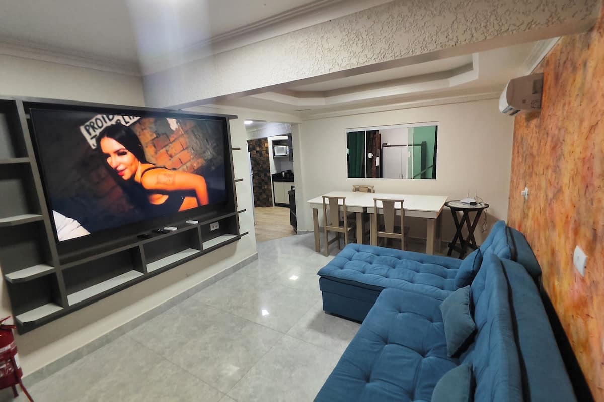 Un salón con televisor de pantalla plana, un sofá azul, una zona de comedor con mesa y sillas, y una cocina al fondo.
