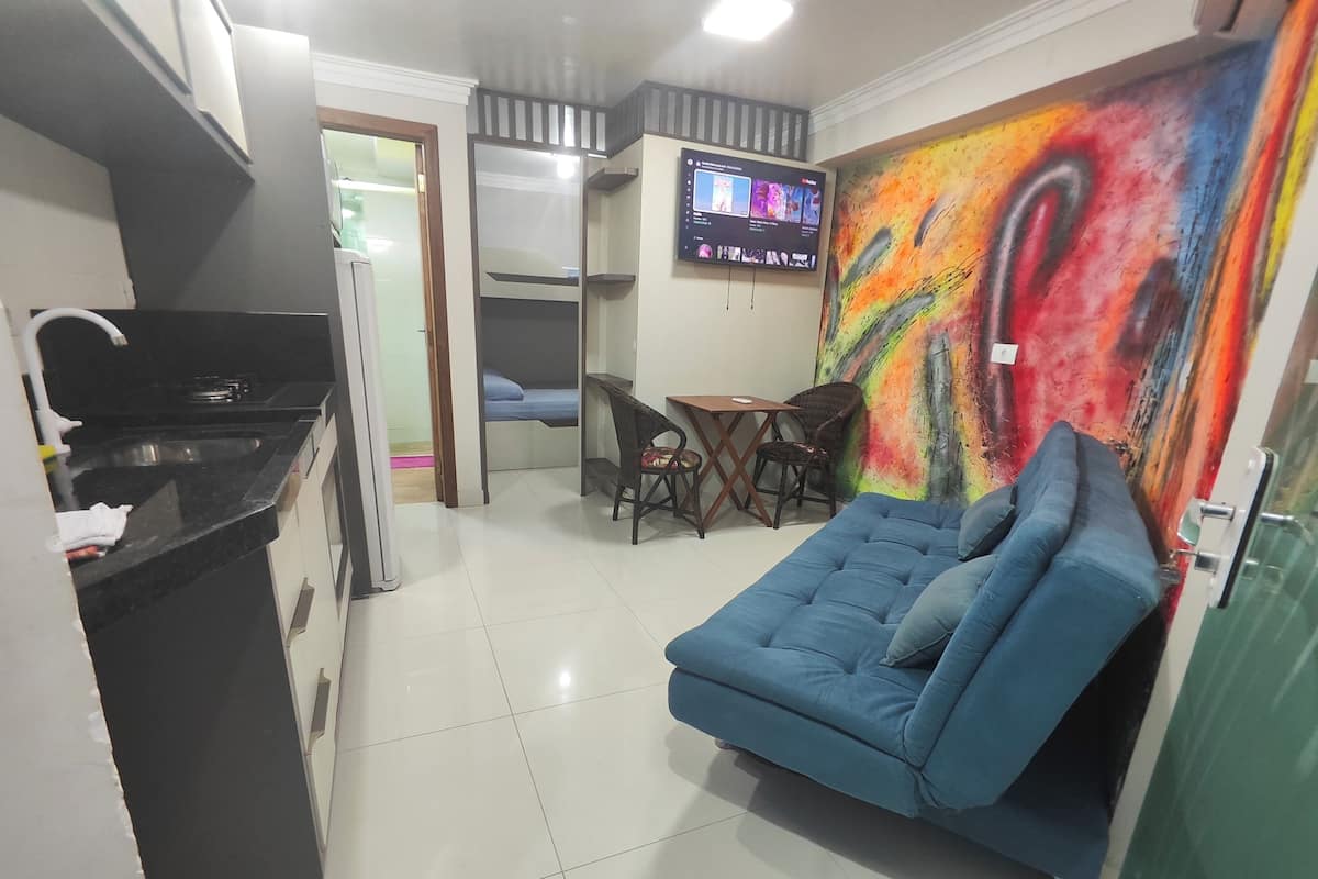 Un espacio habitable compacto con kitchenette, un sofá azul, una mesa de comedor con sillas y un televisor montado en la pared.