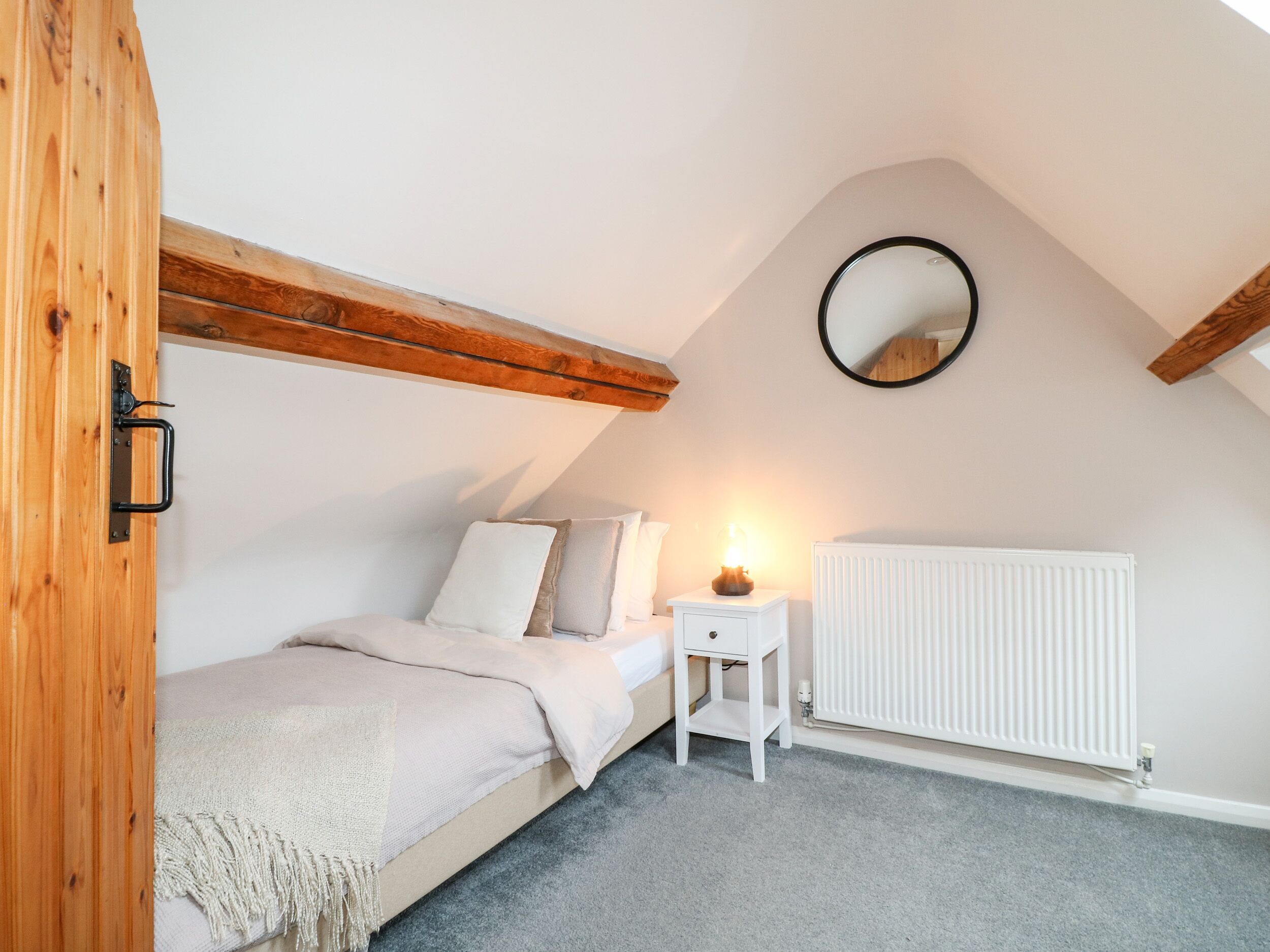 Une chambre avec un plafond en pente, un lit recouvert d’une couverture beige, une table de chevet avec une lampe et un radiateur blanc.
