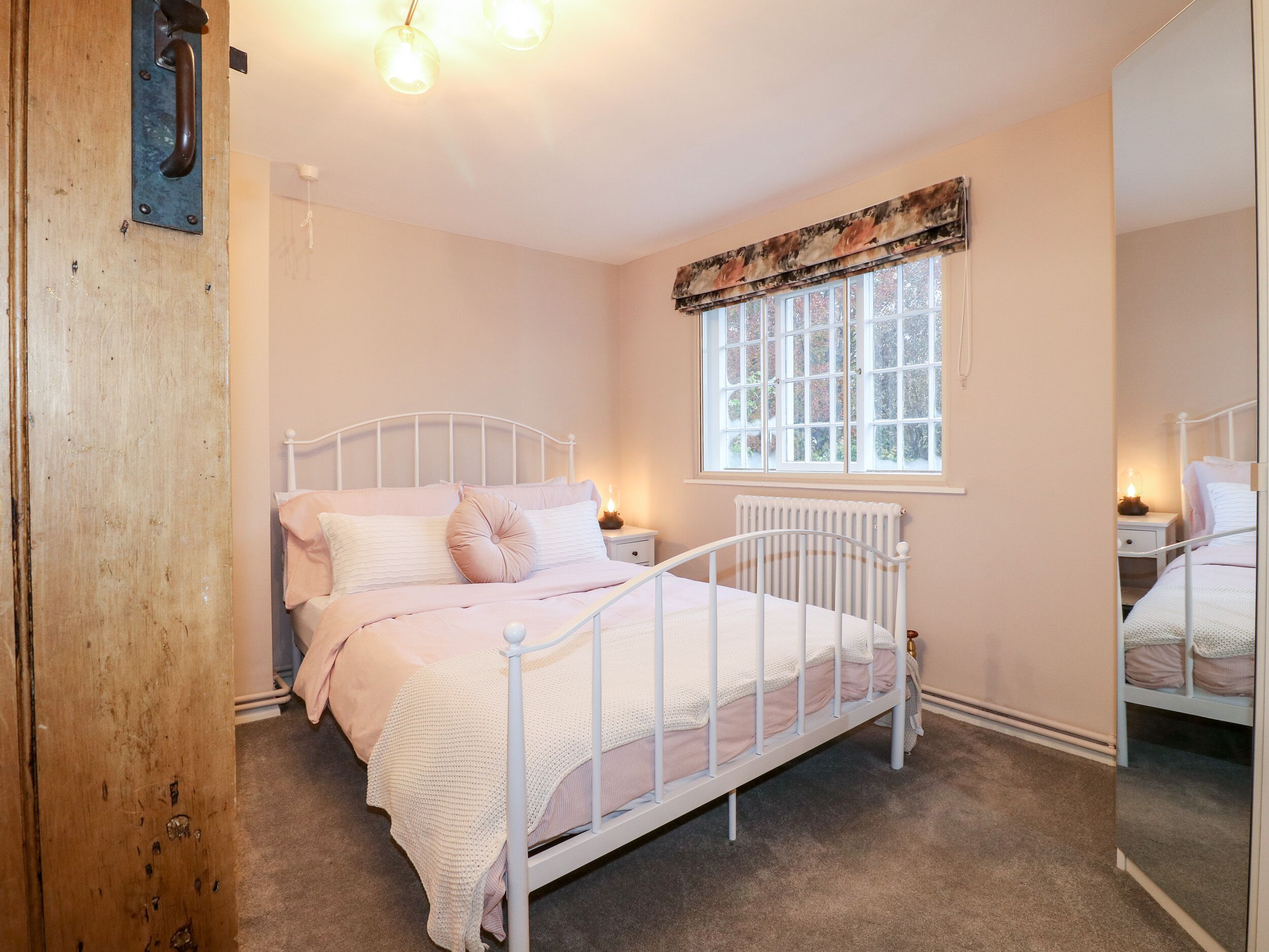 A bedroom with a white metal bed frame, a window with a floral valance, a radiator, and a bedside table with a lamp.