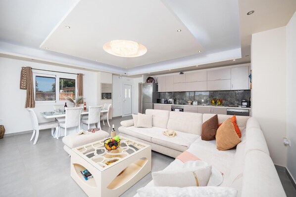 A modern living room with a white sofa, a coffee table, and a dining area with a white table and chairs. - Villa Rudi - Two Bedroom Villa, Sleeps 4 (Ionian Islands)