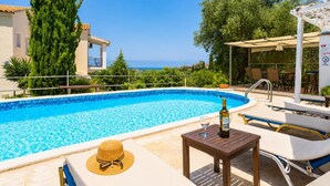 A swimming pool with lounge chairs and a table, a bottle of wine, and a straw hat on the deck. - Villa Mavrikia - Four Bedroom Villa, Sleeps 8 (Sfakera)