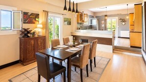 A dining area with a table set for four, a kitchen in the background, and a view of the outdoors through a window. - Villa Mavrikia - Four Bedroom Villa, Sleeps 8 (Sfakera)