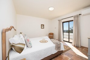 Ein Schlafzimmer mit einem Bett, einer Nachttischlampe, einem Sessel, einem Fenster mit Vorhängen und Blick ins Freie.