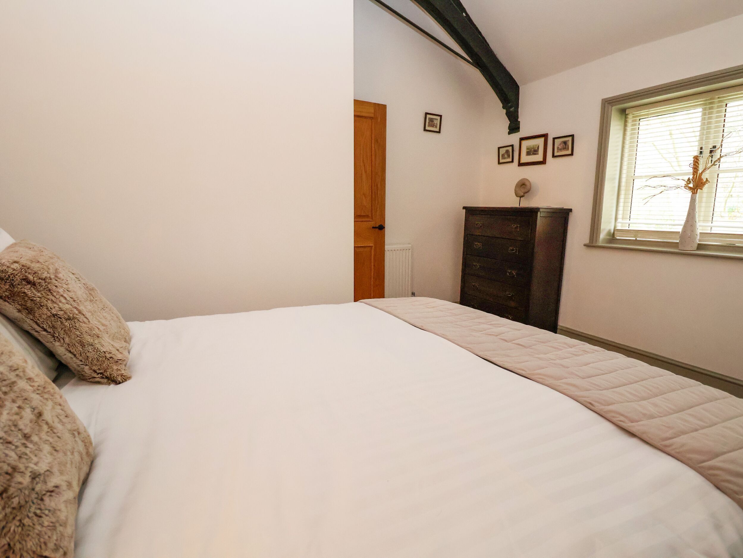 A bedroom with a large bed, a dresser, a window with blinds, and a ceiling with exposed beams.