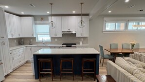 A modern kitchen with white cabinets, a central island with stools, a dining area with a striped sofa, and a large window providing natural light.