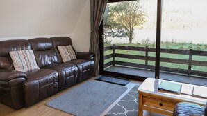 A living room with a brown leather sofa, two pillows, a grey rug, a wooden coffee table, and a view of a wooden deck and trees through large windows. - 27 Invergarry Lodges (Invergarry)