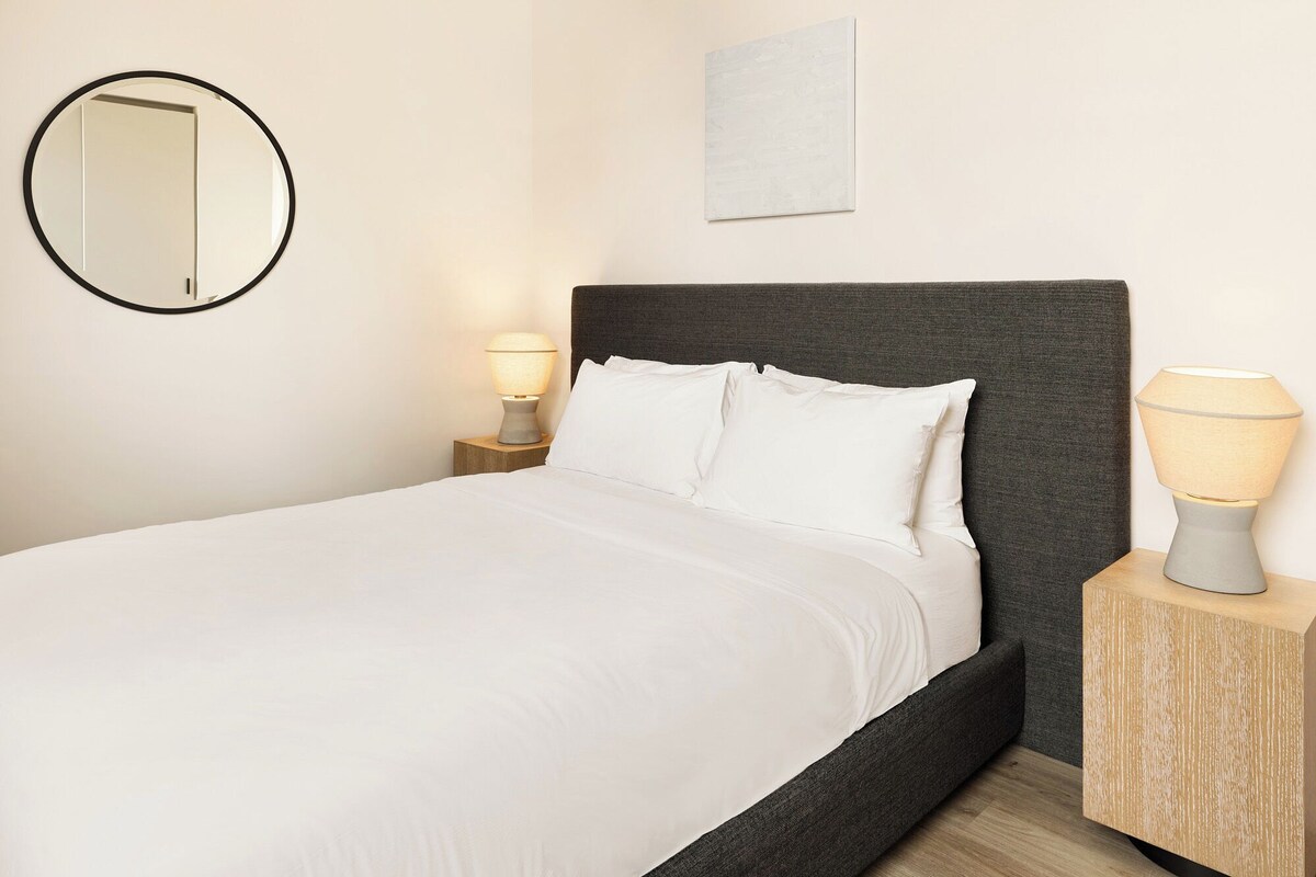 A bed with white bedding, a dark headboard, two bedside lamps, a round mirror, and a small artwork on the wall.