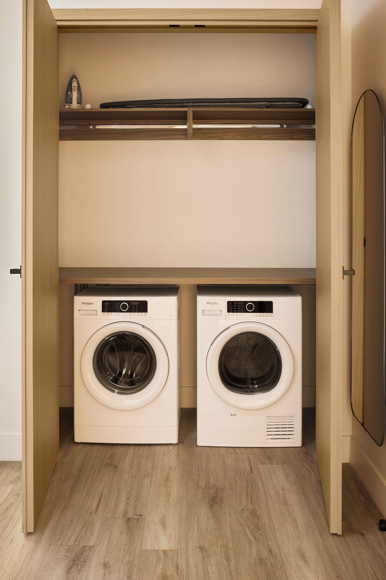 A laundry room with two white Whirlpool washing machines, wooden flooring, and a wooden door.