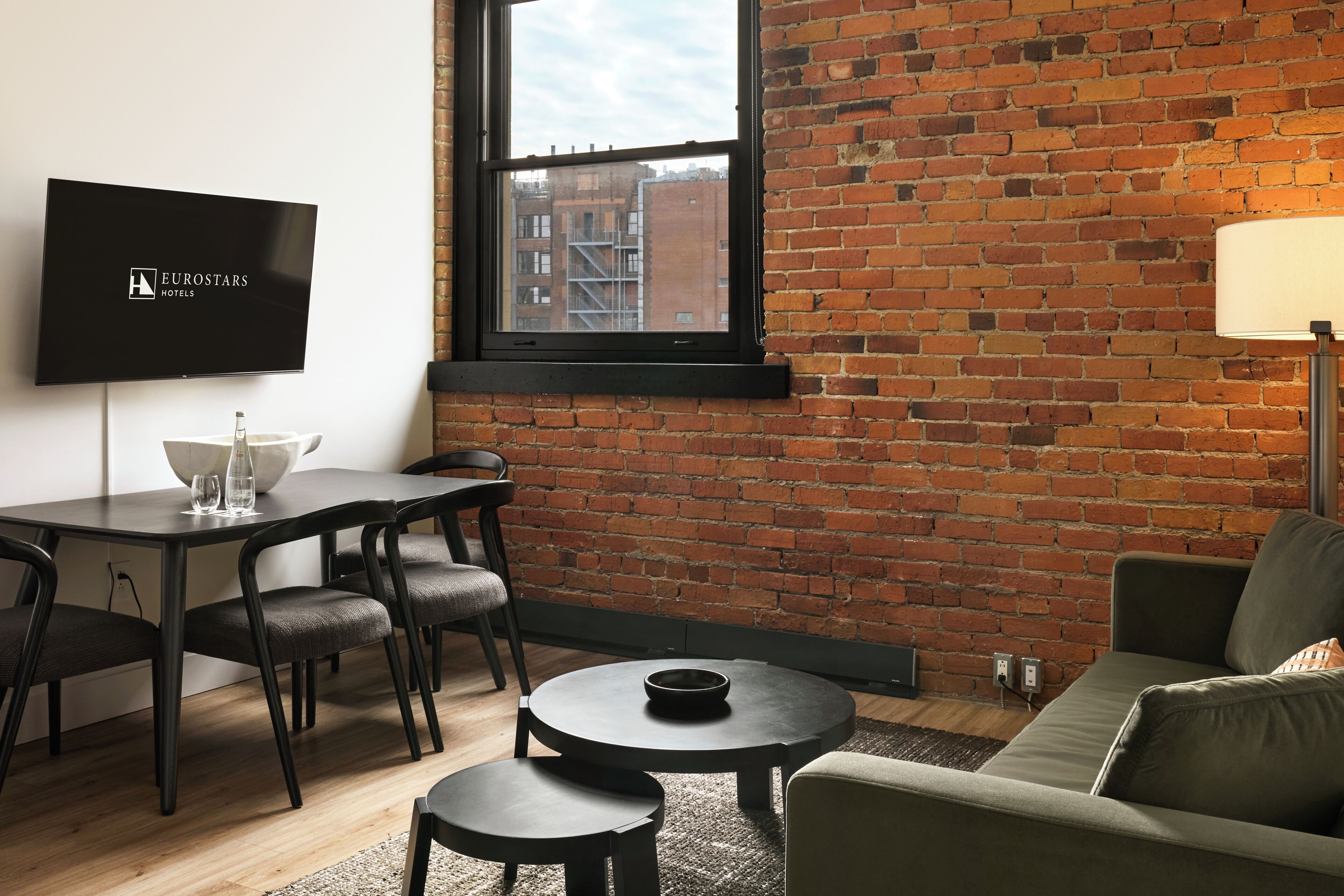 Ein modernes Hotelzimmer mit Backsteinwand, Flachbildfernseher, Essbereich mit Tisch und Stühlen sowie einer Couch.