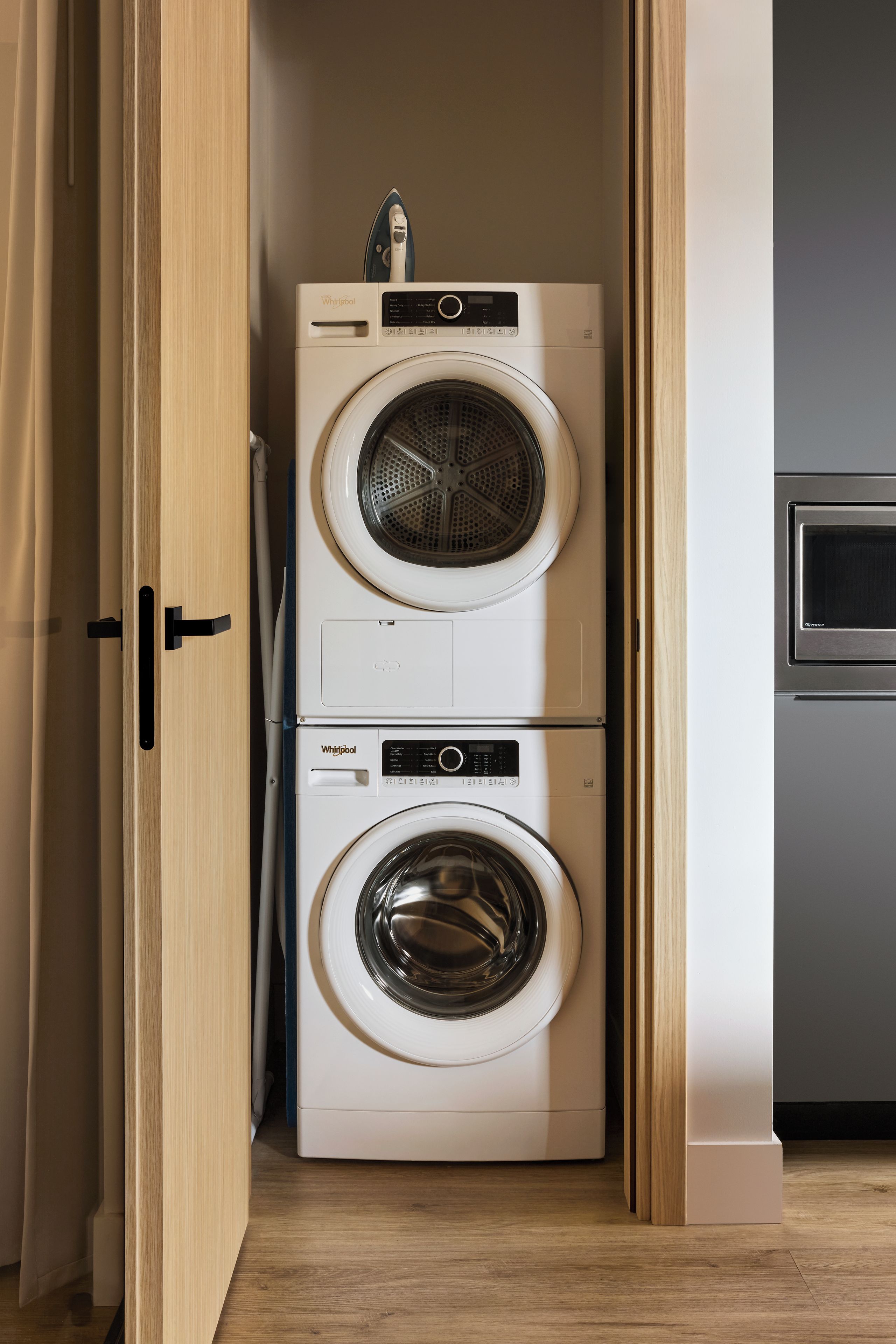 A compact laundry area with two stacked Whirlpool washers and dryers, wooden flooring, and a built-in microwave.