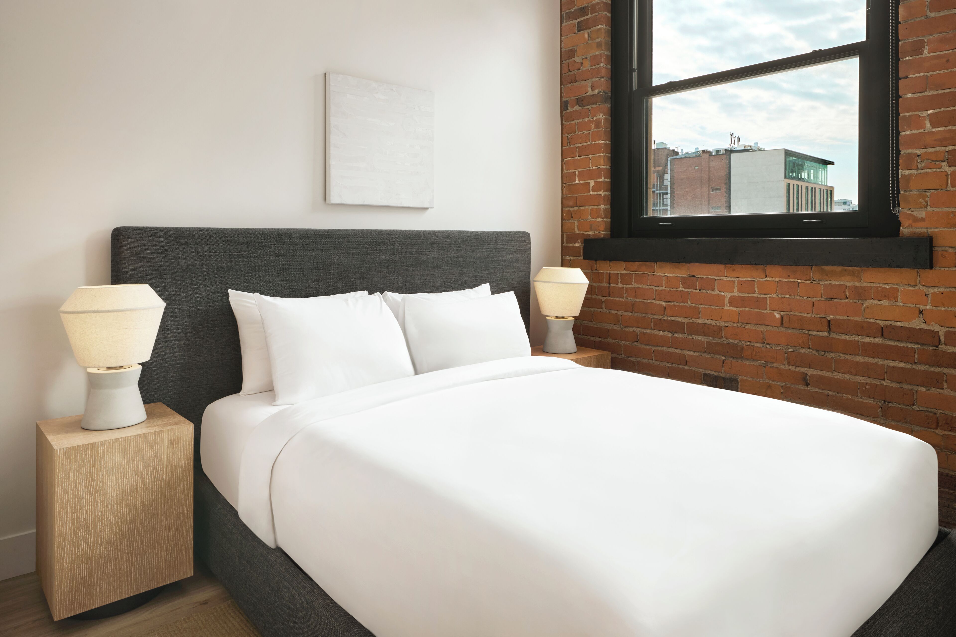A bed with white bedding, two bedside lamps, a wooden nightstand, a brick wall, and a window with a view of buildings.