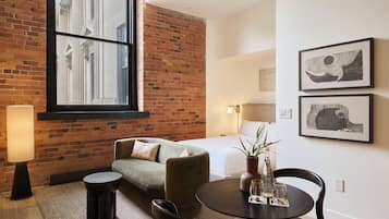 A hotel room with a brick wall, a large window, a sofa, a round table with glasses, and two framed pictures on the wall.