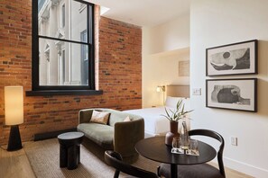 Une chambre d’hôtel avec un mur en briques, une grande fenêtre, un canapé, une table ronde avec des verres et deux tableaux encadrés au mur.