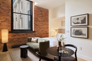 A hotel room with a brick wall, a large window, a sofa, a round table with glasses, and two framed pictures on the wall.