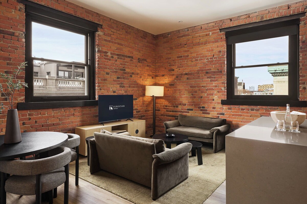 A hotel room with brick walls, a flat-screen TV, a sofa, a small table, and a lamp.