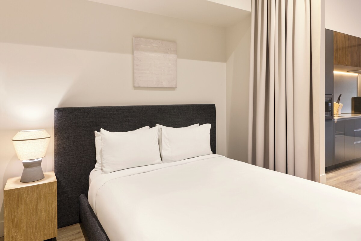 A modern hotel room with a bed, a bedside lamp, a wall-mounted artwork, and a view of the kitchen area.