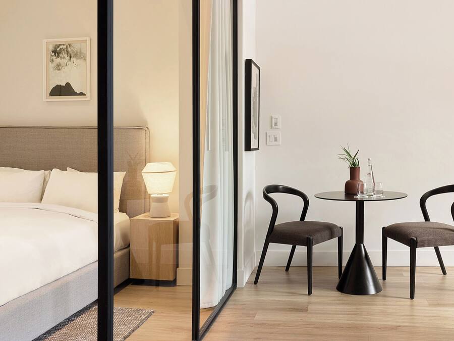 Quarto de hotel com cama, luminária, duas cadeiras, uma mesinha de centro com uma planta e um quadro na parede.