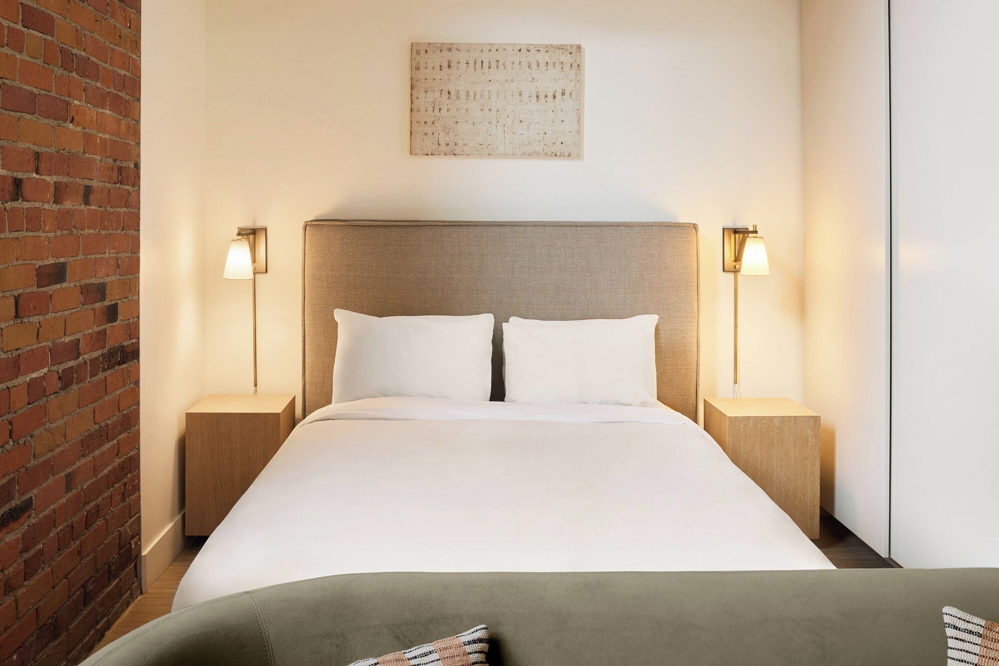 Habitación de hotel con pared de ladrillo, una cama grande con ropa blanca, dos mesitas de noche con lámparas y una obra de arte colgada en la pared.