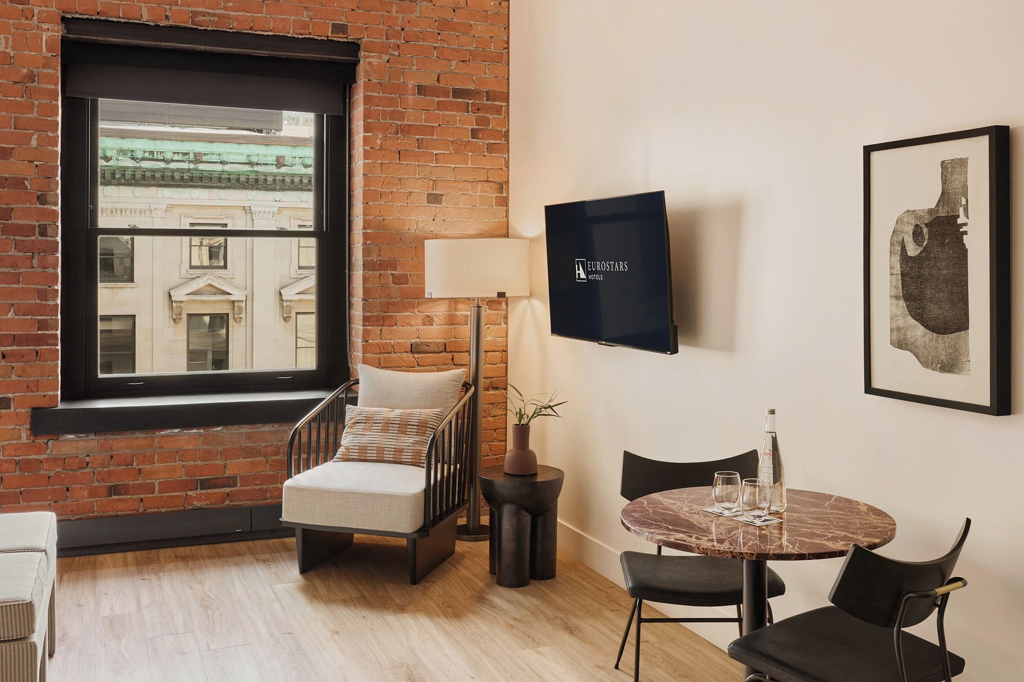A hotel room with a brick wall, a large window, a bed, a chair, a table with chairs, a TV, and artwork.