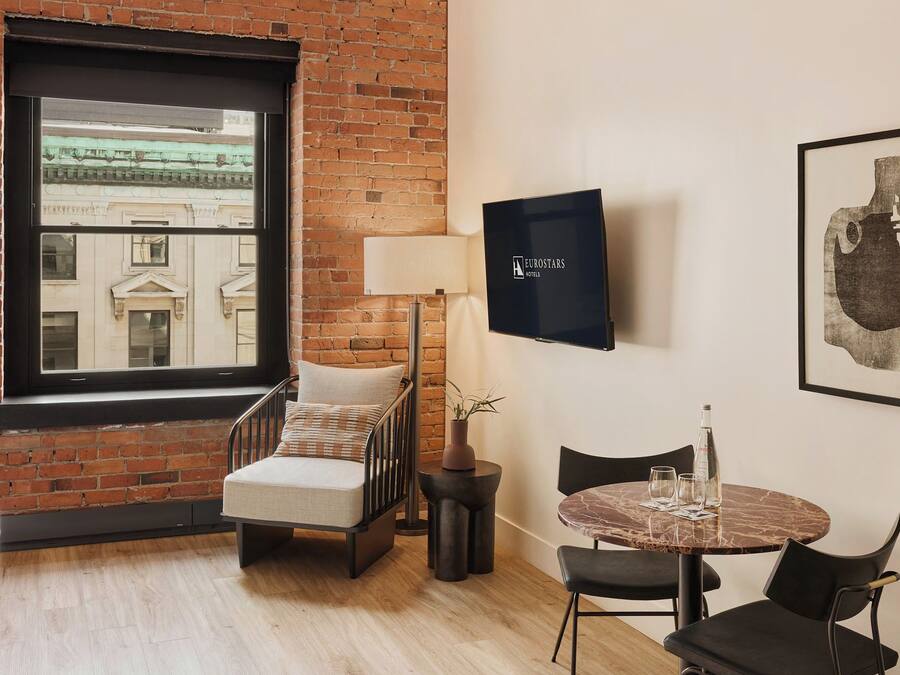 A hotel room with a brick wall, a large window, a bed, a chair, a table with chairs, a TV, and artwork.