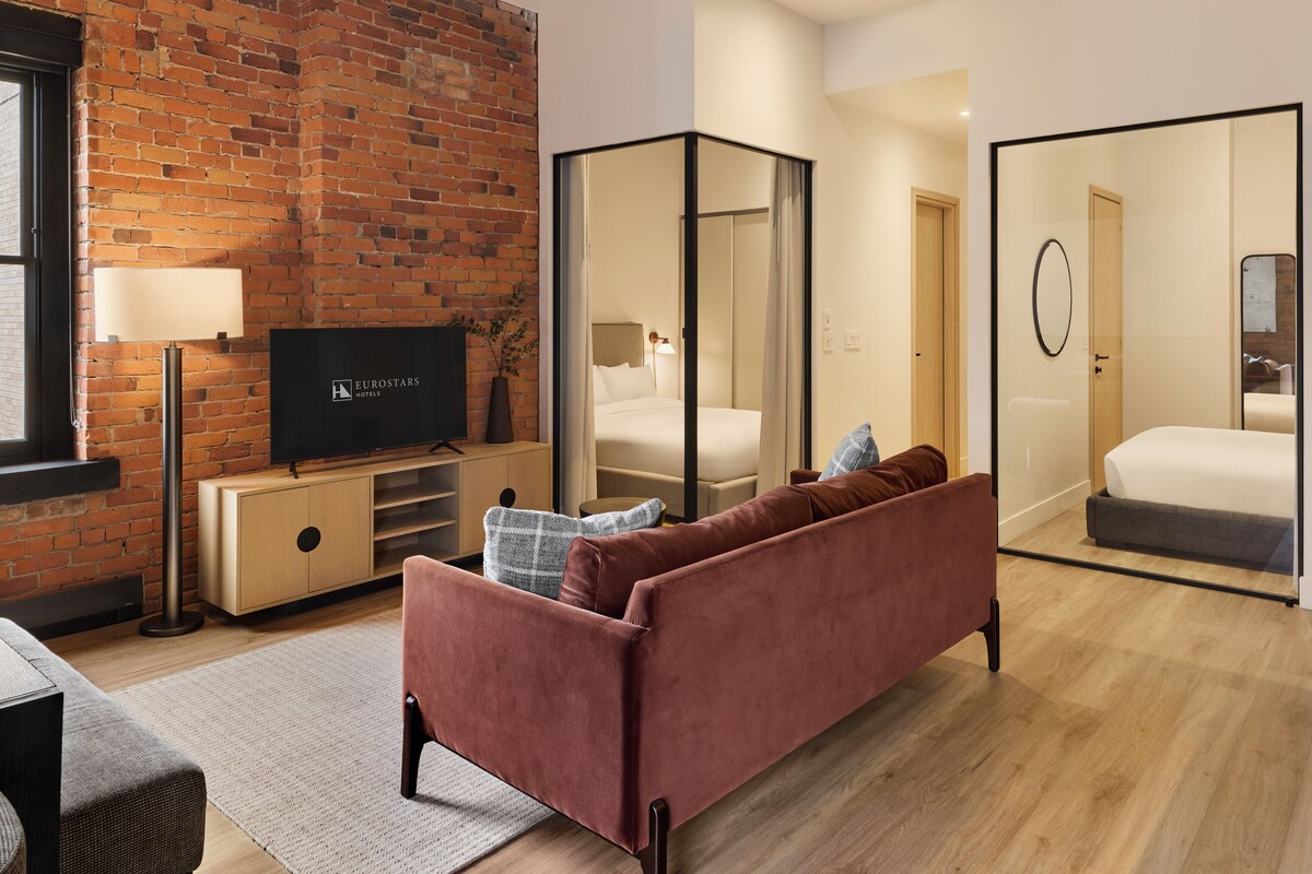 A modern hotel room with a brick wall, a flat-screen TV, a sofa, a bed, and a mirror.
