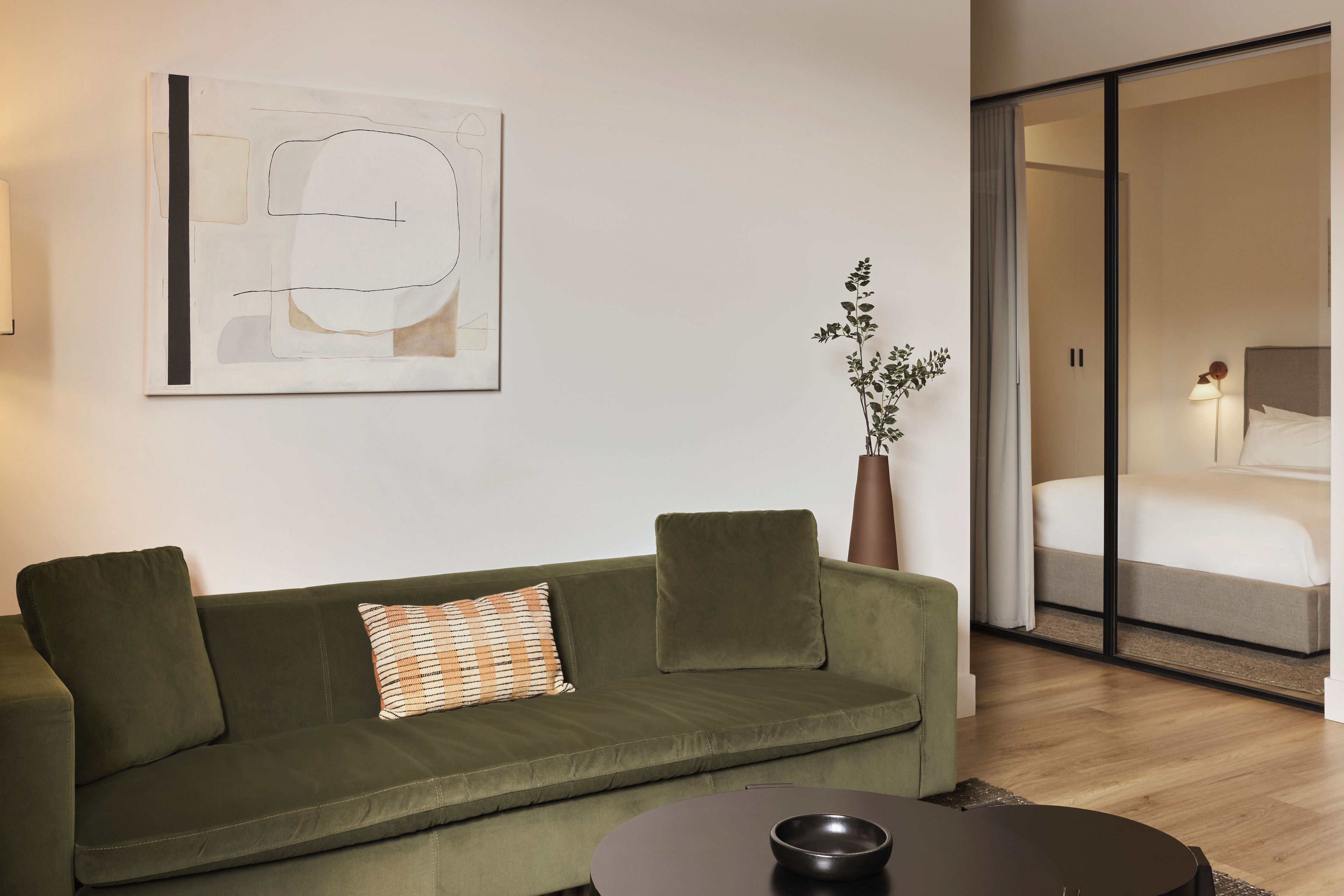 A modern hotel room with a green sofa, a single bed, a large window, and a minimalist abstract painting on the wall.