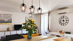 Una cocina moderna con una isla central, luces colgantes, una zona de comedor con mesa y sillas, un televisor y un reloj de pared.