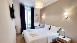 Una cama bien hecha con sábanas y toallas blancas, una mesita de noche con lámpara, y vistas al exterior a través de una ventana con cortinas.