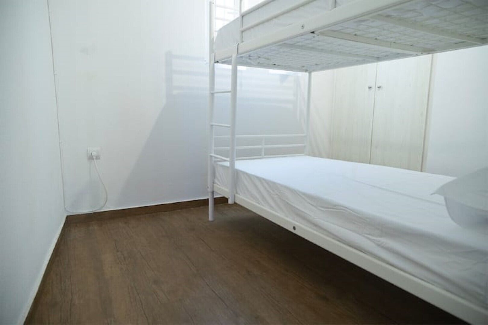 A small, single-bed room with a bunk bed, wooden flooring, white walls, and a visible electrical outlet.