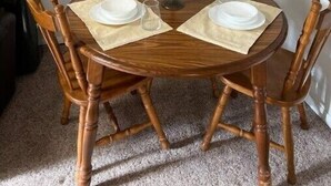 Een ronde houten eettafel met vier stoelen, een vaas met bloemen en een lijst met een afbeelding aan de muur.