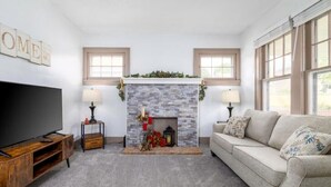 A living room with a stone fireplace, a television, a sofa, and two windows. - The Cedar House (Steubenville)