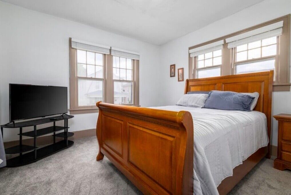 Une chambre à coucher avec un lit en bois, un téléviseur posé sur un meuble et de grandes fenêtres équipées de stores.