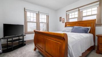 Une chambre à coucher avec un lit en bois, un téléviseur posé sur un meuble et de grandes fenêtres équipées de stores.