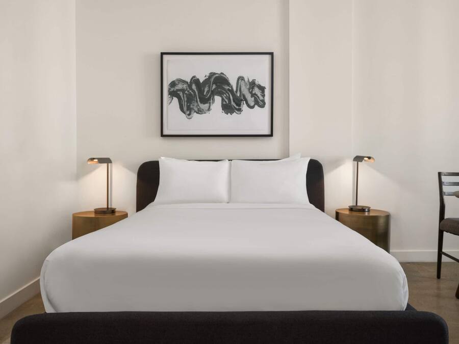 A neatly made bed with white linens, two bedside tables with lamps, a framed artwork on the wall, and a chair to the right.