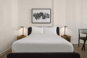 A neatly made bed with white linens, two bedside tables with lamps, a framed artwork on the wall, and a chair to the right. - Reside Craftsman Los Angeles, a Wyndham Residence (Los Angeles)