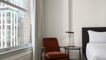 A hotel room with a bed, a chair, a side table, and a window with a view of a building.