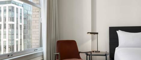 A hotel room with a bed, a chair, a side table, and a window with a view of a building.