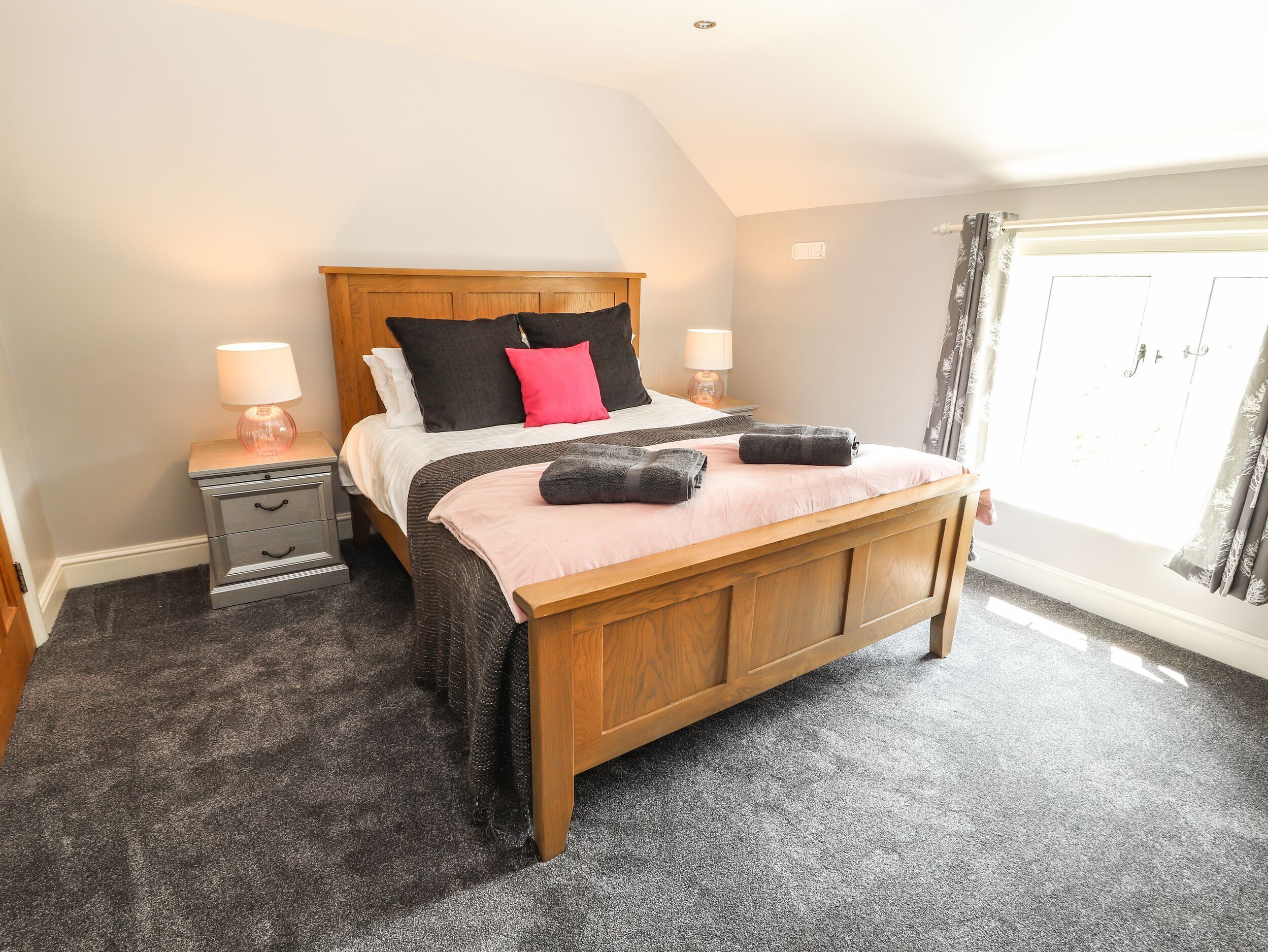 A bedroom with a wooden bed, two bedside tables with lamps, a large window with curtains, and a carpeted floor.