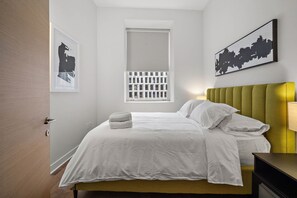 Un lit bien fait, avec du linge de lit blanc, une tête de lit recouverte d’un tissu vert et une fenêtre avec des stores.